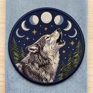 May include: A circular embroidered patch featuring a gray and white wolf howling toward a series of moon phases and gold stars against a dark blue sky, framed by green pine trees. The design is stitched with detailed thread work.