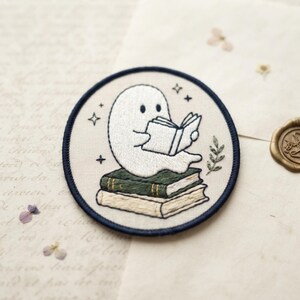 May include: A circular embroidered patch featuring a white ghost sitting on a stack of two books while reading an open book. The design includes small black stars and a green leafy sprig on a cream background with a dark blue stitched border.