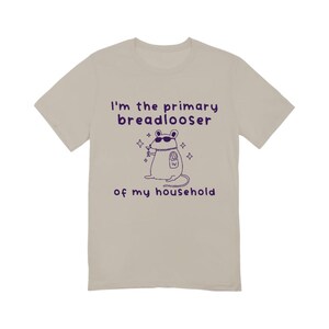 May include: A beige short-sleeve t-shirt featuring a dark purple graphic of a mouse wearing sunglasses and a chain, holding a drink. The text reads: I'm the primary breadlooser of my household. The design includes small star accents around the mouse.