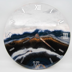 May include: Circular wall clock with a resin design in white, navy blue, and gold hues. The clock face has silver Roman numerals and hands. The design evokes a layered landscape effect.