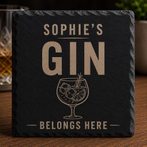 May include: A square black slate coaster with rough, chiseled edges. The surface features light-colored engraved text that reads SOPHIE'S GIN BELONGS HERE, accompanied by a line-art illustration of a stemmed glass containing ice and a lime slice.