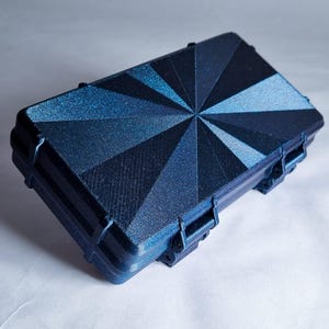 Può includere: Una valigetta rettangolare blu navy con un design geometrico sul coperchio. Il coperchio presenta un motivo radiale di forme triangolari in varie tonalità di blu. La valigetta ha una superficie strutturata e chiusure sicure.