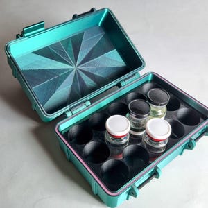 May include: An open teal case with a geometric pattern on the inside lid. The case holds small glass jars with black and white lids. The case is designed to securely hold the jars, ideal for storage.