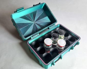 May include: An open teal case with a geometric pattern on the inside lid. The case holds small glass jars with black and white lids. The case is designed to securely hold the jars, ideal for storage.