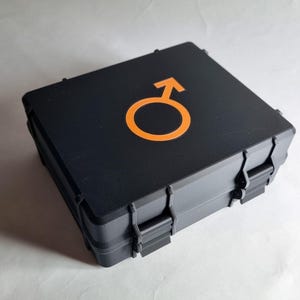 May include: A black, rectangular case featuring a bright orange Mars symbol. The case has a textured surface and secure latches. The Mars symbol is a circle with an arrow pointing upwards and to the right.