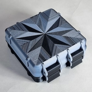 Könnte beinhalten: Eine quadratische zweifarbige Box mit einem Sternmuster auf dem Deckel. Der Deckel ist hellblau-grau mit einem schwarzen Sternmuster. Die Box hat schwarze Verschlüsse und eine hellblau-graue Basis.