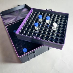 Könnte beinhalten: Eine schwarze und lila Aufbewahrungsbox mit einem Raster aus kreisförmigen Löchern, die mehrere kleine Glasfläschchen mit blauen und weißen Kappen enthält. Die Box ist geöffnet und zeigt das Innere und den Deckel mit einem geometrischen Muster.