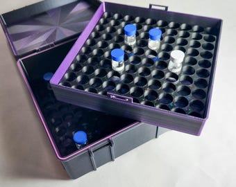 Könnte beinhalten: Eine schwarze und lila Aufbewahrungsbox mit einem Raster aus kreisförmigen Löchern, die mehrere kleine Glasfläschchen mit blauen und weißen Kappen enthält. Die Box ist geöffnet und zeigt das Innere und den Deckel mit einem geometrischen Muster.