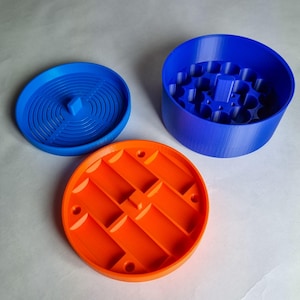 Könnte beinhalten: Dreiteiliges Set aus 3D-gedruckten Kunststoffkomponenten in Blau und Orange. Das Set umfasst einen zylindrischen Behälter, einen Deckel mit konzentrischen Rillen und eine Basis mit geometrischem Muster. Die Teile sind separat angeordnet.
