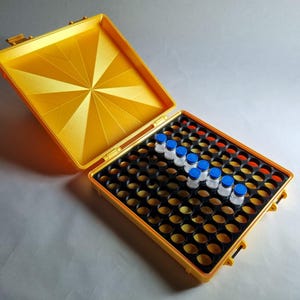 Op de afbeelding: Open gele koffer met een geometrisch ontwerp op het deksel, met meerdere kleine flesjes met blauwe doppen. De koffer heeft een zwart interieur met talrijke cirkelvormige compartimenten. De flesjes zijn in een rij gerangschikt.