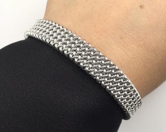May include: A silver-colored bracelet with a woven mesh design. The bracelet is worn on a wrist, and the background is a black fabric.