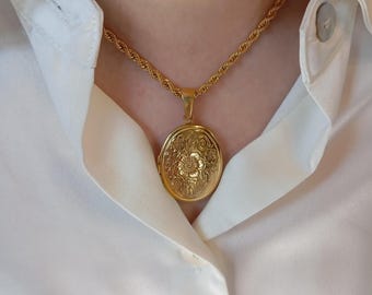 May include: A gold-colored locket necklace with a floral design pendant. The pendant hangs from a twisted rope chain. The necklace is worn over a white collared shirt.