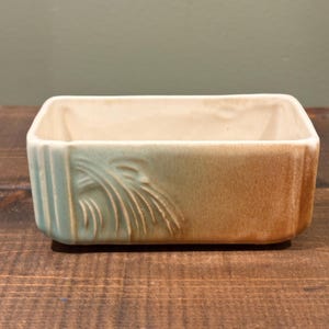 May include: Rectangular ceramic dish with a two-tone color scheme. The interior is a light beige, while the exterior transitions from a pale turquoise to a warm brown. The side features a stylized, carved leaf design, adding a touch of elegance.