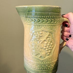 May include: A vintage ceramic pitcher with a light green and cream color scheme. The pitcher features a detailed embossed grape design and decorative bands. The handle is curved, and the spout is angled for pouring.