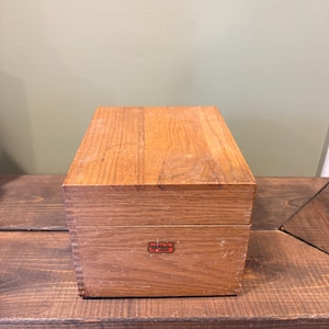 May include: A rectangular wooden box with a natural wood grain finish. The box has a small, red and white emblem on the front. The box is sitting on a dark wooden surface.