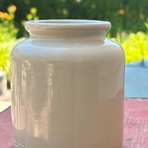 May include: A small, cylindrical off-white ceramic jar with a smooth, glossy finish. The vessel features a short, slightly flared rim at the top and a flat base. The surface is plain and uniform, showing a subtle embossed mark near the bottom edge.