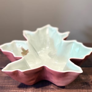 May include: A ceramic bowl shaped like a maple leaf with a pale mint green interior and a matte dusty rose pink exterior. The bowl features a small gold-colored decorative accent on the inner surface, mimicking the appearance of a small insect.