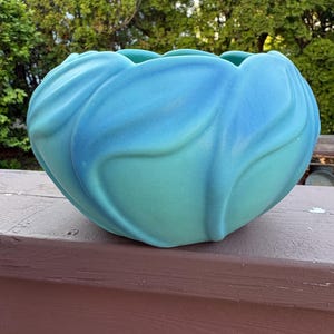 May include: A large, round, ceramic bowl in shades of blue and teal. The bowl has a textured, petal-like design around the exterior. The bowl is sitting on a brown wooden surface, with a green background of trees.