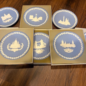May include: A collection of blue and white decorative plates, each featuring a different iconic London landmark. The plates are displayed in their original boxes, with a white laurel wreath border. The plates are approximately 6 inches in diameter.