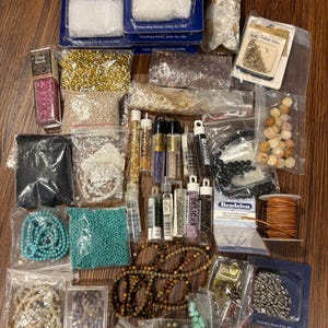May include: Assortment of beads and jewelry making supplies in various colors and materials, including glass beads, metal beads, gemstone beads, and cord. The supplies are packaged in small plastic bags and containers.