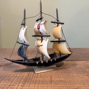 May include: A miniature wooden sailboat with three masts and tan sails. The sails are adorned with a red and white emblem and the word "Lugano". The boat is black with a small stand.