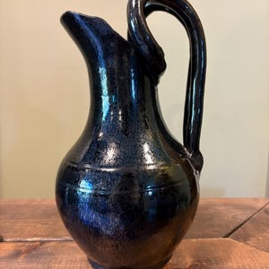 May include: A dark blue and black glazed ceramic pitcher with a rounded body, a flared spout, and a tall, curved handle that loops from the rim to the shoulder of the vessel. The surface features a mottled, reflective finish.