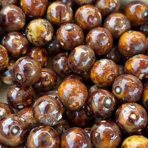 Pode incluir: Um close-up de uma coleção de contas de cerâmica marrom e branco. As contas são redondas e têm um acabamento brilhante. Elas estão dispostas em um padrão aleatório.