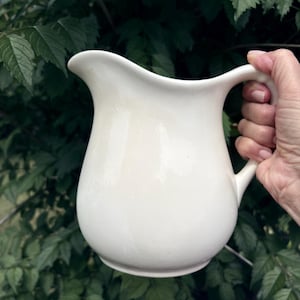 May include: A solid off-white ceramic pitcher with a smooth, glossy finish, featuring a curved handle on the right side and a wide, rounded pouring spout at the top. The vessel has a bulbous body that tapers slightly toward the base.