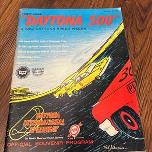 Puede incluir: Un programa de recuerdo vintage para el Daytona 500 de 1962, con una vibrante ilustración de coches de carreras en una pista. La portada es predominantemente azul, roja y amarilla, con el texto "Daytona 500" y "Daytona International Speedway."