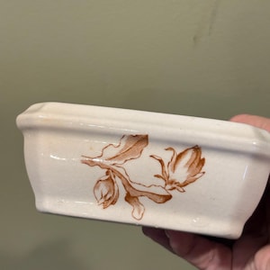 May include: A rectangular off-white ceramic dish with a beveled rim and a glossy finish. The exterior features a brown botanical illustration of a flower bud and leaves on a stem. The ceramic surface shows a subtle crackle texture throughout.