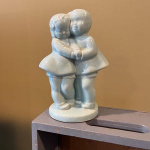 May include: A ceramic figurine of two children hugging, painted in a light blue glaze.