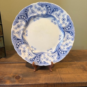 May include: A white ceramic plate with a blue floral design. The plate has a scalloped edge and is supported by a small wooden stand. The blue pattern includes flowers and swirling lines.