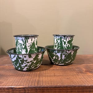 May include: Two sets of enamelware bowls and cups. Each set includes a small cup nested inside a larger bowl. The bowls and cups have a white base with a green splatter pattern and black rims. The items are displayed on a wooden surface.