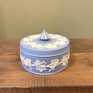 May include: A round, light blue trinket box with a white relief design of grape vines and leaves. The lid has a conical finial and matching relief details. The box sits on a wooden surface.