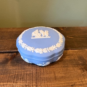 May include: A light blue ceramic trinket box with a white relief design. The lid features a central scene of figures and is framed by a white floral border. The box is round and sits on a wooden surface.