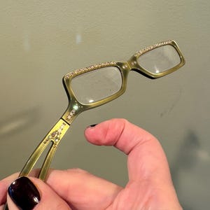 May include: A pair of gold-toned eyeglasses with rectangular lenses. The frames are embellished with small, sparkling rhinestones along the top and at the hinges. The glasses are held in a hand with dark nail polish.