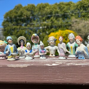 May include: A collection of 11 vintage porcelain doll heads with painted details. The dolls are wearing various hats and dresses in shades of blue, pink, yellow, and white.