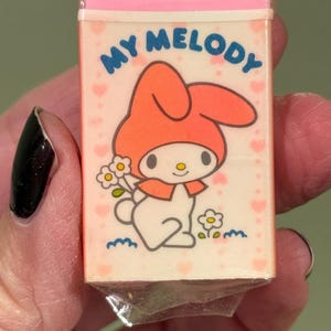 May include: A rectangular eraser with a pink top and a My Melody graphic. The graphic features a white bunny wearing an orange hood, holding flowers, and the words "MY MELODY" in blue.