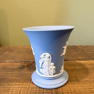 May include: A light blue ceramic vase with white relief detailing. The vase features classical figures and floral designs. The vase has a flared top and a circular base, and is set on a wooden surface.
