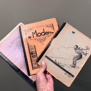 May include: Three vintage softcover booklets with black tape spines. The left booklet is light purple with the text Le Calligraphe. The center orange booklet features the text le Moderne. The right tan booklet shows a skier and the text LE SKI.