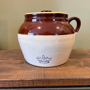 May include: A vintage brown and white stoneware crock with a lid and handle. The crock features a dark brown top and a white base with a blue emblem. This antique kitchenware is perfect for farmhouse decor.