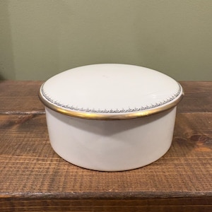 May include: A white ceramic round box with a lid. The lid and base are white, with a gold-colored band around the rim. The box sits on a wooden surface.
