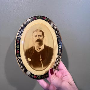 May include: Oval-shaped picture frame with a black and white checkered border and a floral design. The frame contains a black and white photograph of a man with a mustache.
