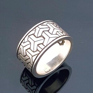 May include: A silver ring with a geometric pattern. The ring has a wide band with a repeating design of interlocking shapes. The ring is highly reflective, and the design is visible on the ring's surface.