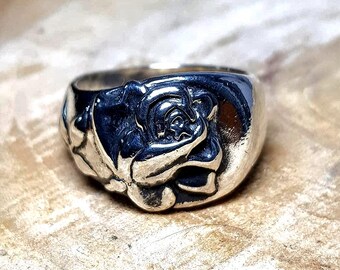 May include: A silver ring with a detailed rose design. The ring features a raised floral motif on a wide band, showcasing the petals and leaves. The ring is made of a shiny, reflective metal.
