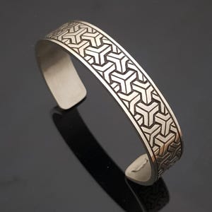 May include: Silver-toned cuff bracelet with a geometric pattern. The open cuff design features a repeating pattern of interlocking shapes across the top surface. The bracelet is made of a shiny, metallic material.