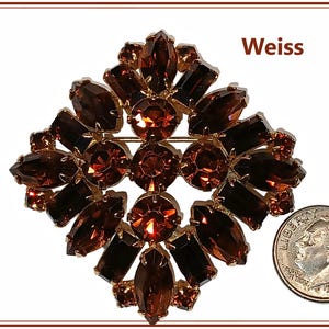 May include: A vintage Weiss brooch featuring a diamond-shaped design with brown and amber-colored rhinestones set in a gold-tone metal. The brooch has a pin closure on the back. A dime is included for size reference.