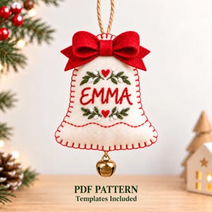 May include: A cream-colored felt bell ornament featuring a red felt bow at the top, red blanket stitching along the edges, and the name EMMA embroidered in red thread. The design includes green leaf and red heart embroidery, with a gold metal bell.