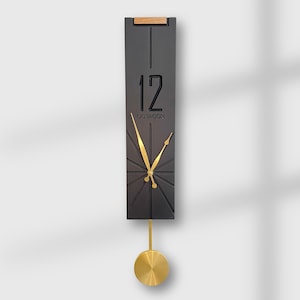 Puede incluir: Un reloj de pared rectangular moderno con una esfera gris oscuro y manecillas y péndulo dorados. El reloj muestra el número 12 y la palabra "DOTAGON". La parte superior del reloj tiene un acento de madera.