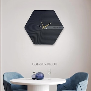 May include: A black, hexagonal wall clock with gold hands and the text "OQTAGON". The clock face features concentric circular lines. The text "OQTAGON DECOR" is below the clock. The clock is mounted on a white wall.